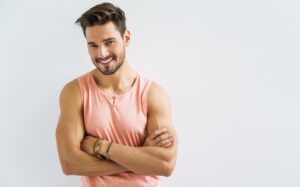 A man smiling at the camera and crossing his arms, while wearing a pink tank top.