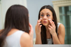 A woman pulling at and examining her under eye bags in the mirror
