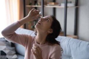 A woman sitting on a bed and putting eyedrops in her eye