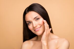 A woman smiling at the camera while tilting her face and touching her cheek